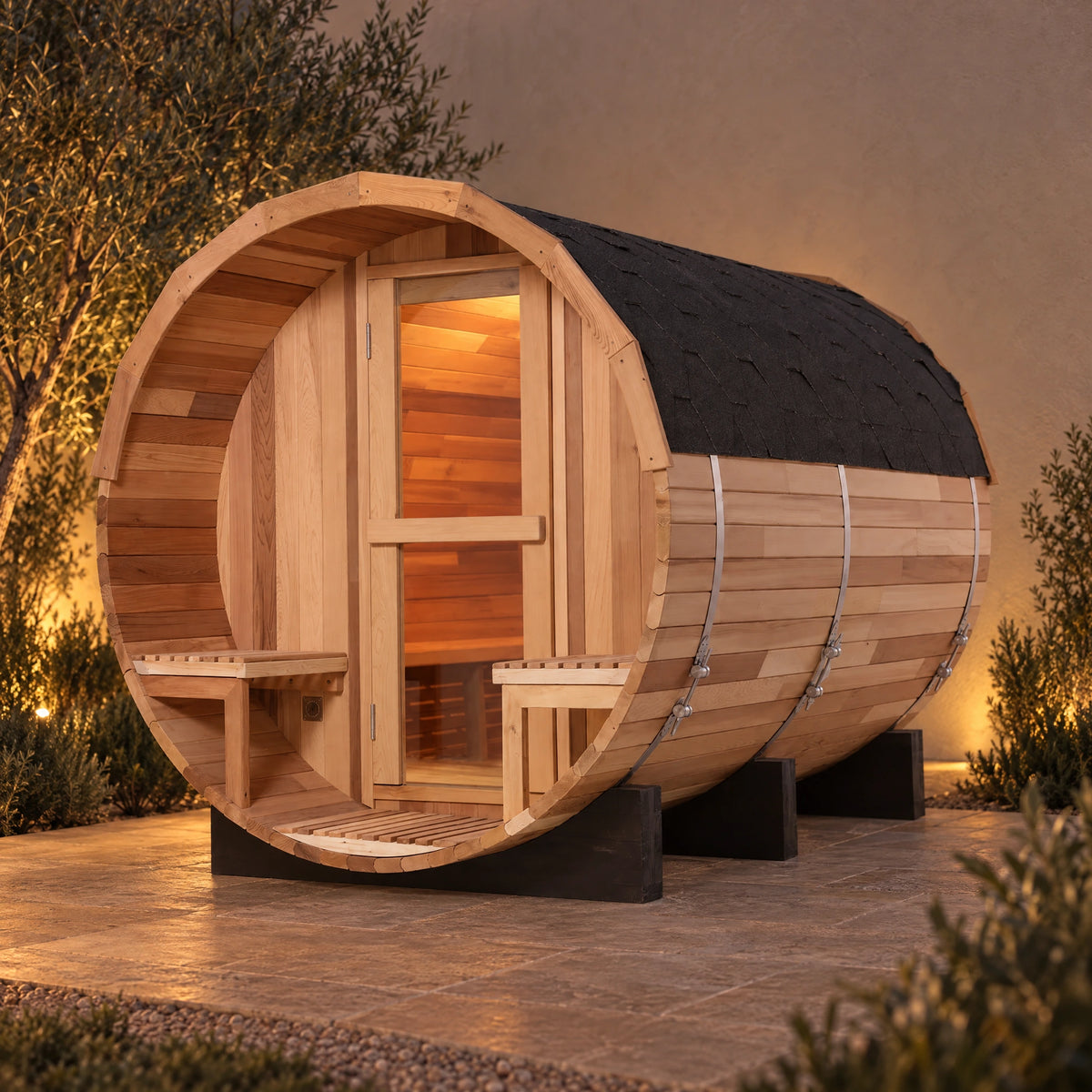 Evening main view of the cedar barrel sauna on a quiet stone terrace, with warm light through the glass entrance.